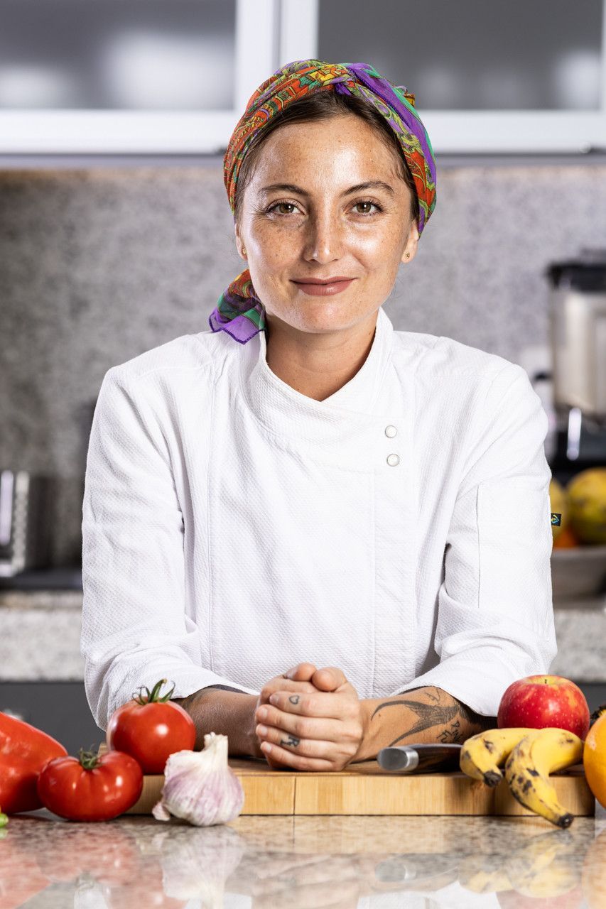 Chef de cozinha de jaleco branco sorrindo em uma cozinha com tomates, alho, bananas e uma tábua de cortar.