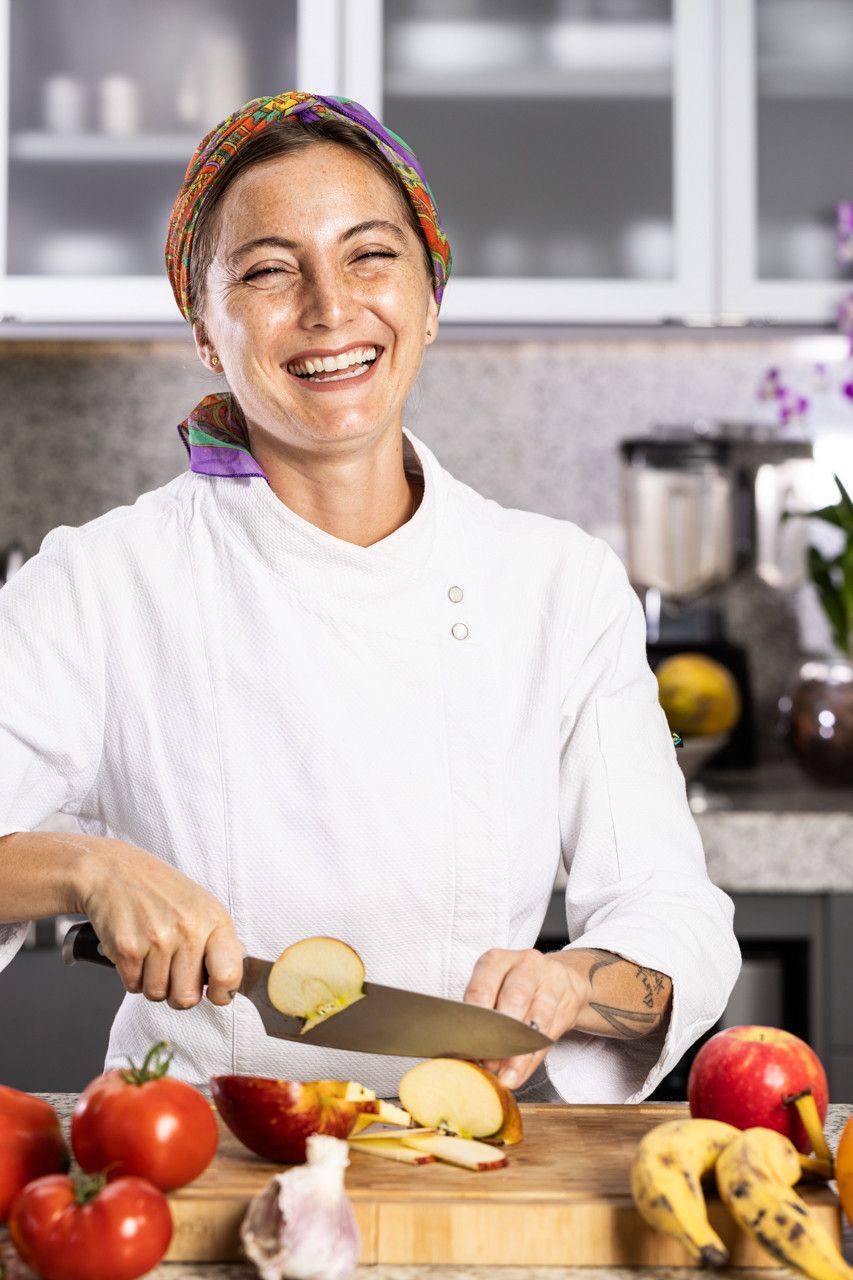 Chef sorridente cortando batatas em uma cozinha com tomates, bananas e um liquidificador na bancada.