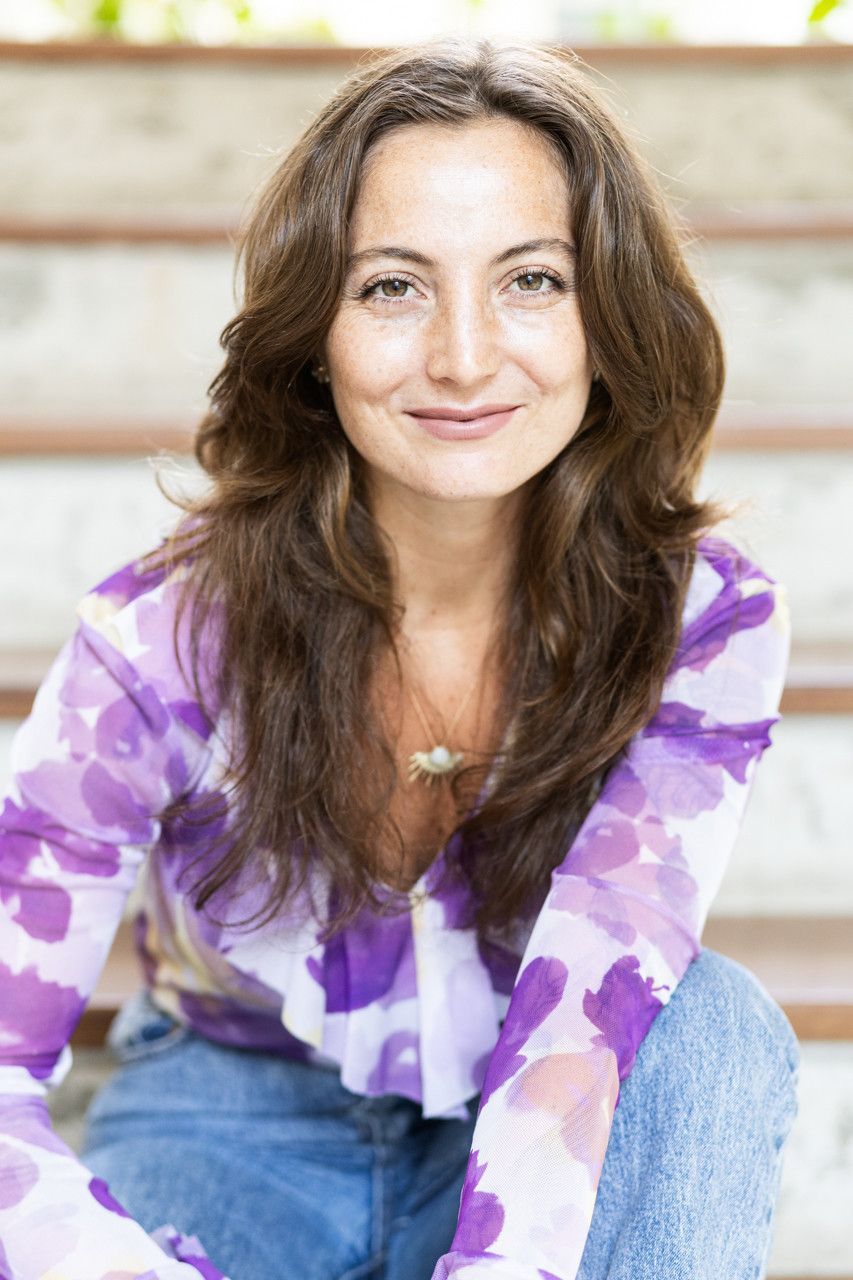 Mulher com blusa floral roxa e calça jeans, sorrindo enquanto está sentada em uma escada ao ar livre.