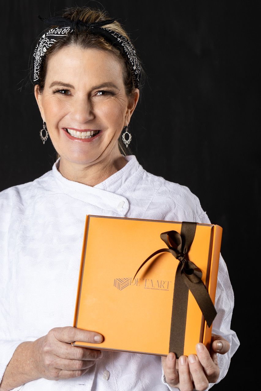 Uma pessoa sorridente vestida de branco segura uma caixa de presente laranja amarrada com uma fita preta.