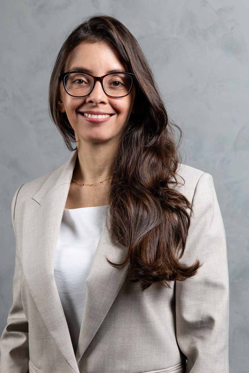 Pessoa sorridente com longos cabelos escuros e óculos, vestindo um blazer claro, contra um fundo cinza.