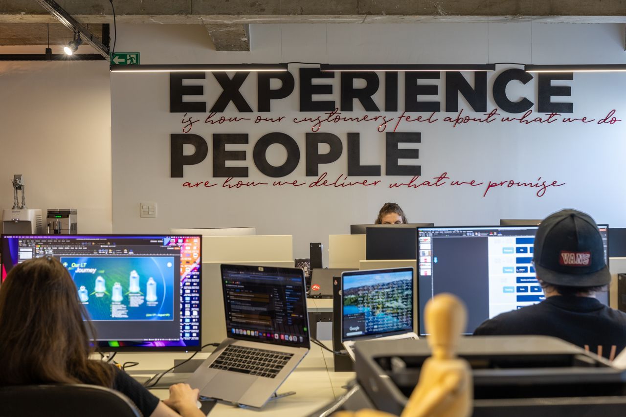 Pessoas trabalhando em computadores em um escritório moderno sob uma placa na parede que diz