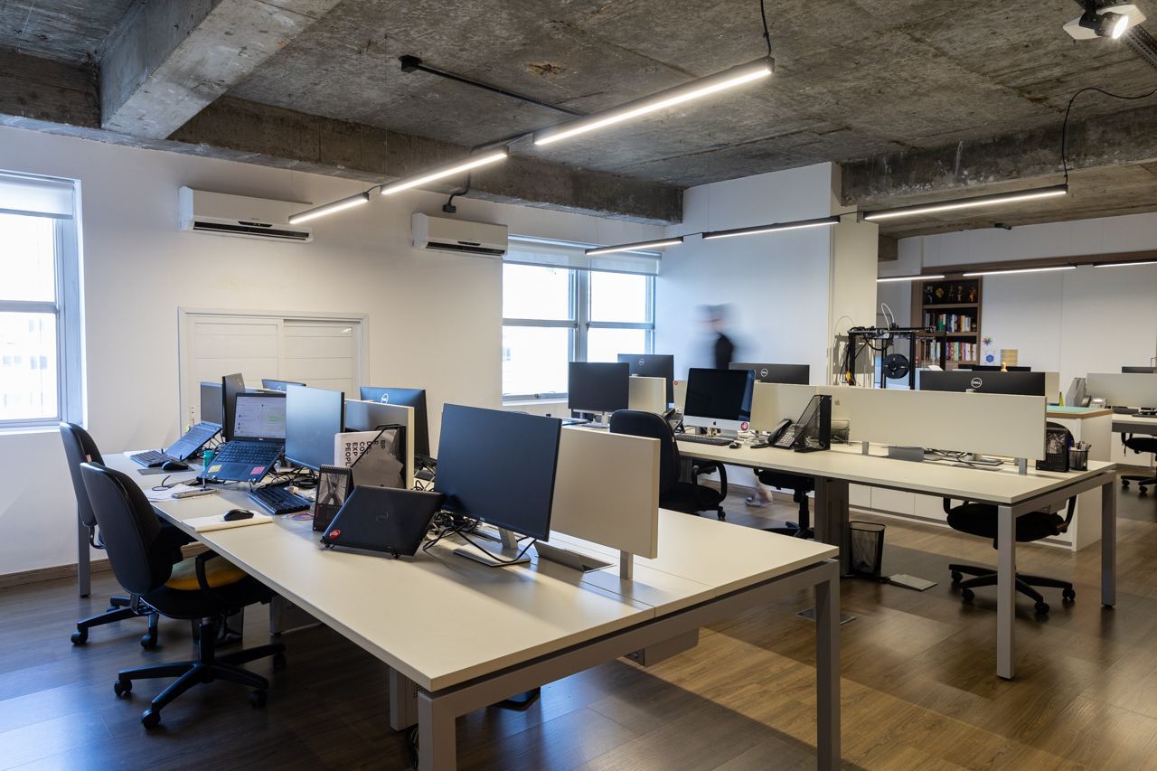 Escritório em plano aberto com mesas, monitores de computador, cadeiras e grandes janelas sob iluminação aparente no teto.