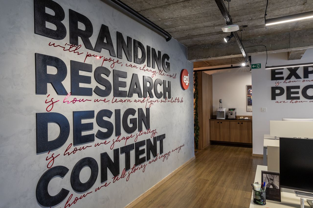 Corredor de escritórios com um mural cinza na parede com os dizeres “BRANDING RESEARCH DESIGN CONTENT” e uma recepção nas proximidades.