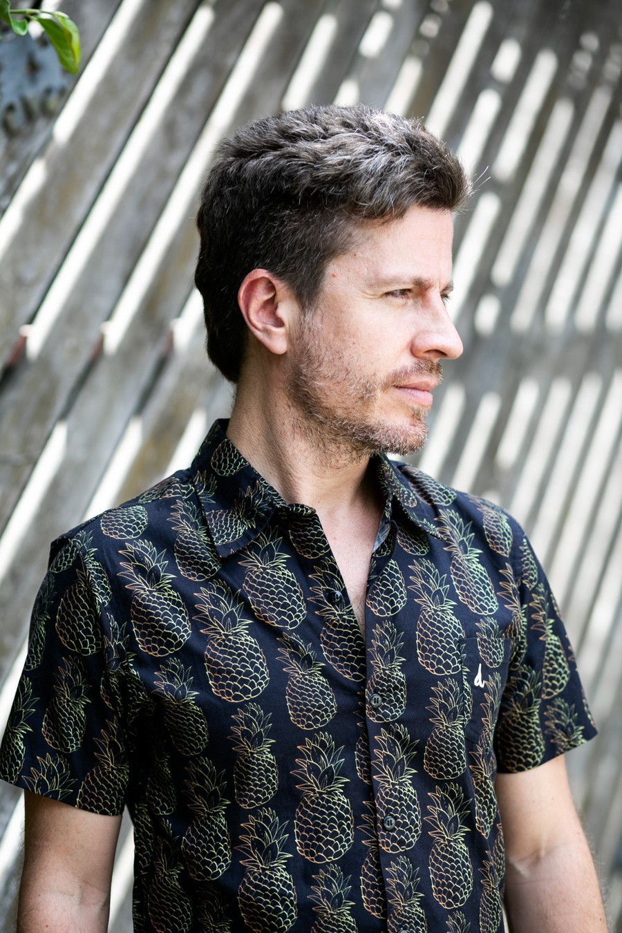 Homem de pé ao ar livre, vestindo uma camisa escura estampada, olhando para o lado contra uma parede listrada.
