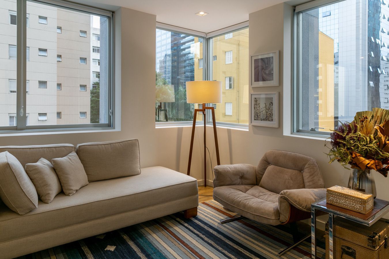 Sala de estar moderna com sofá bege, poltronas, tapete listrado, luminária de chão e amplas janelas com vista para a cidade.