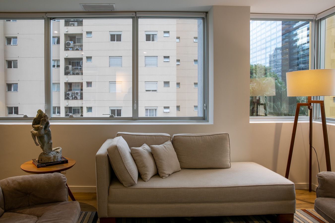Sala de estar moderna com sofá bege, luminária de chão e amplas janelas com vista para prédios de apartamentos.