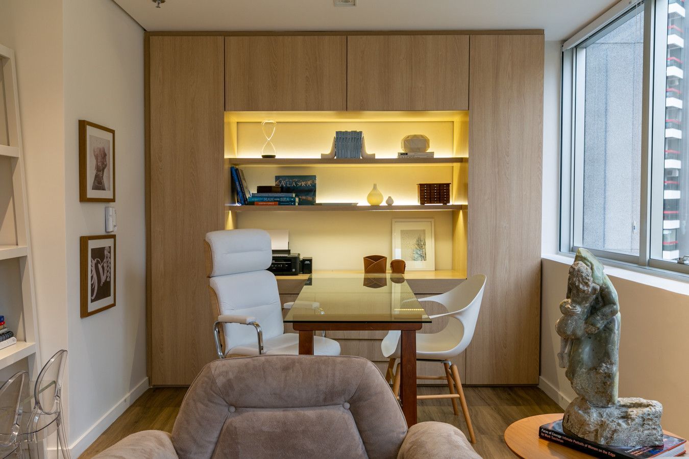Escritório moderno em casa com mesa, cadeira branca, prateleiras embutidas, iluminação aconchegante e parede com painéis de madeira.