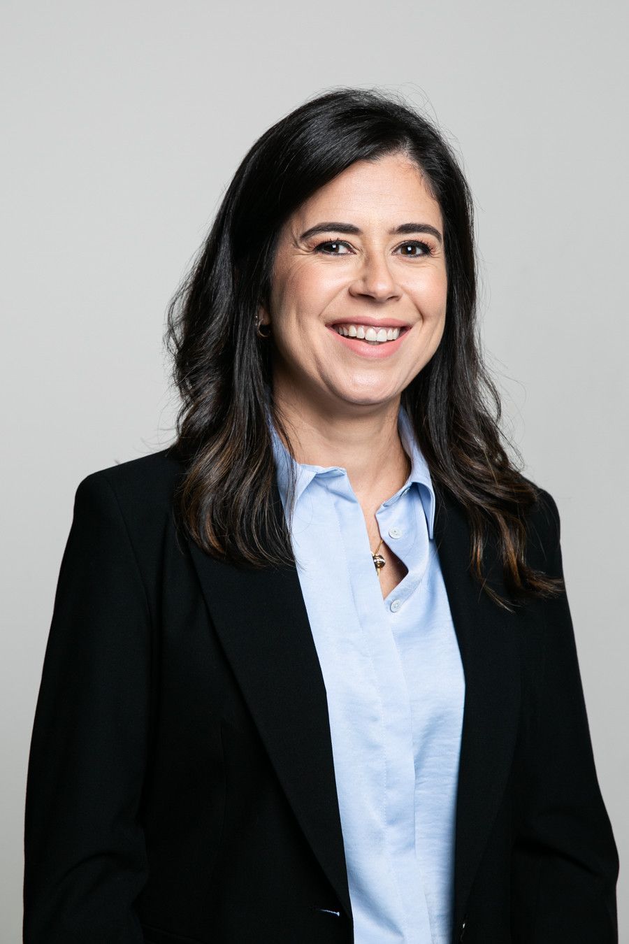 Mulher sorridente com blazer preto e camisa azul-clara contra um fundo cinza.