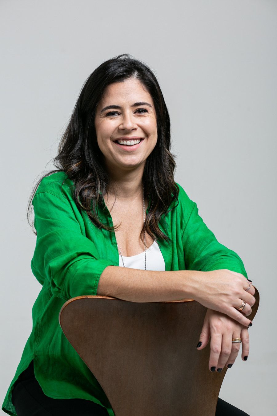 Mulher de jaqueta verde sorrindo enquanto se apoia em uma cadeira contra um fundo cinza.