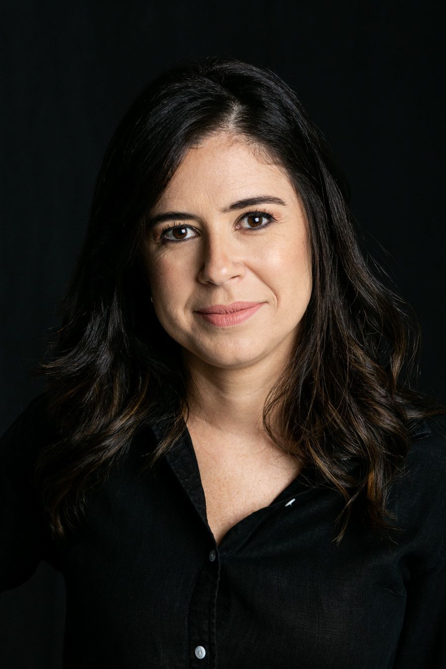 Retrato de estúdio de uma mulher de cabelos escuros vestindo uma camisa preta contra um fundo escuro.