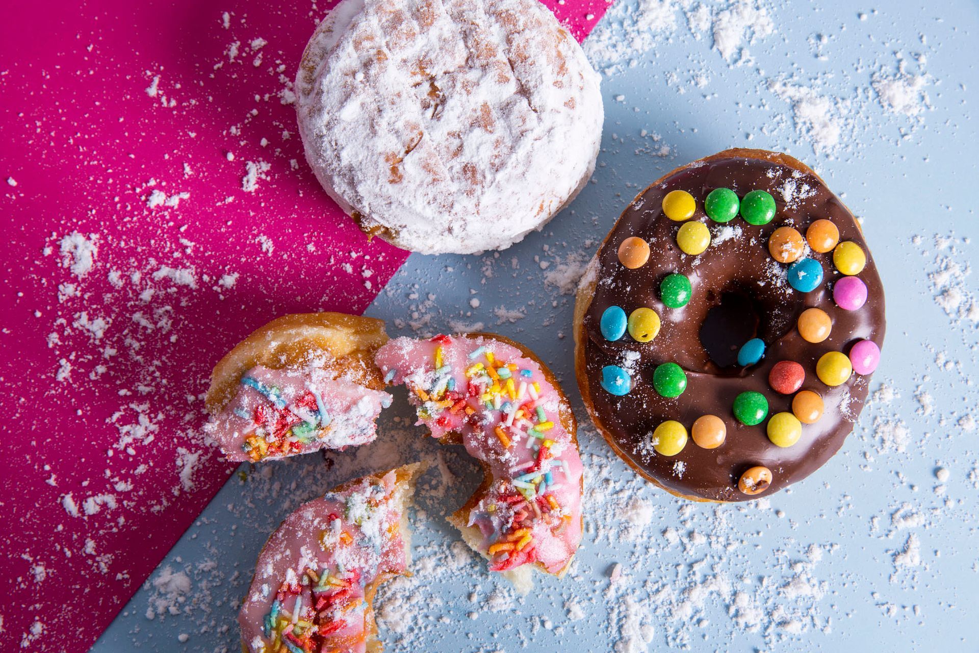 Três donuts sobre fundo rosa e azul: um polvilhado com açúcar, um de chocolate com pedaços de doce e um mordido e com granulado.
