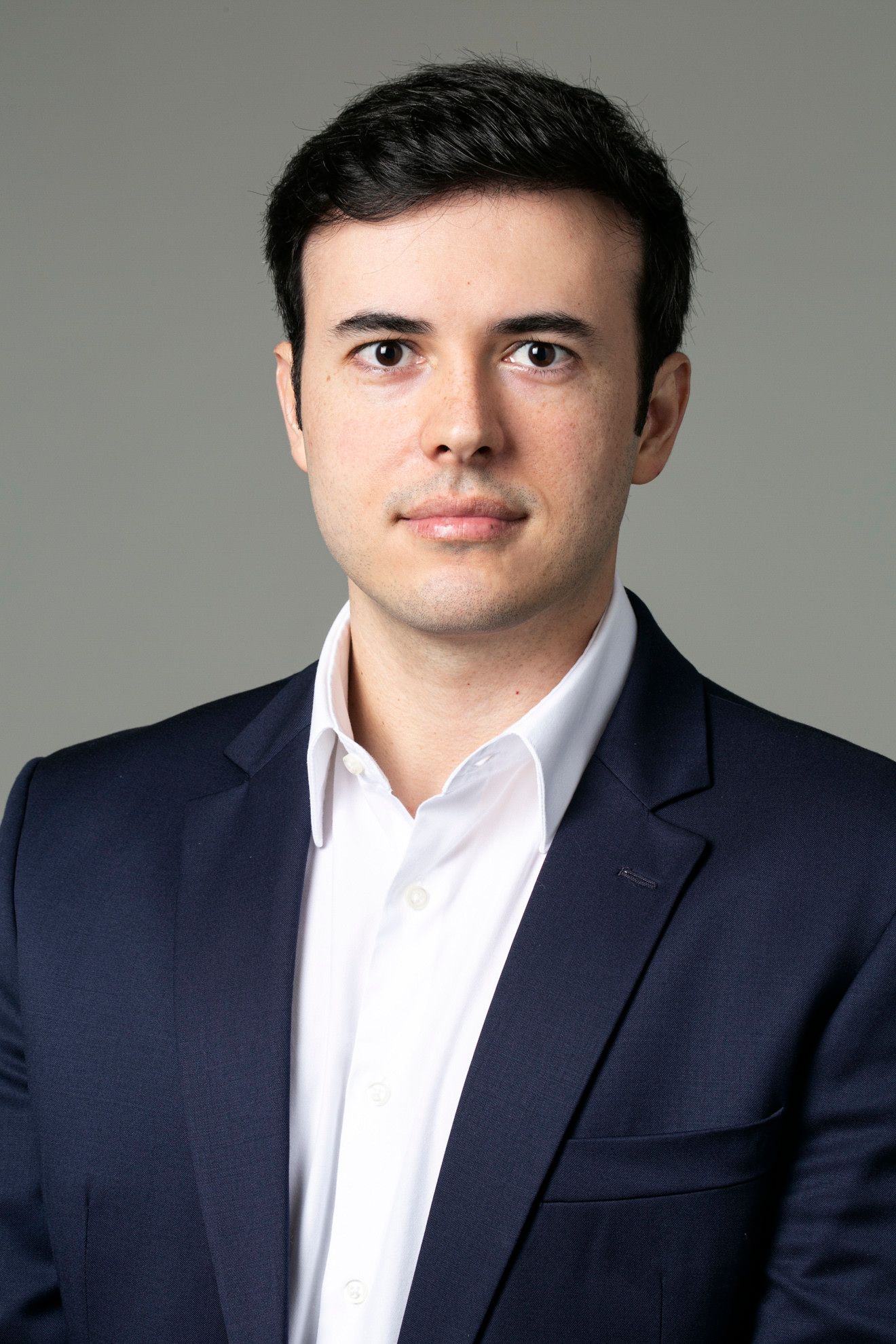 Retrato de estúdio de um homem de terno azul-marinho e camisa branca contra um fundo cinza.