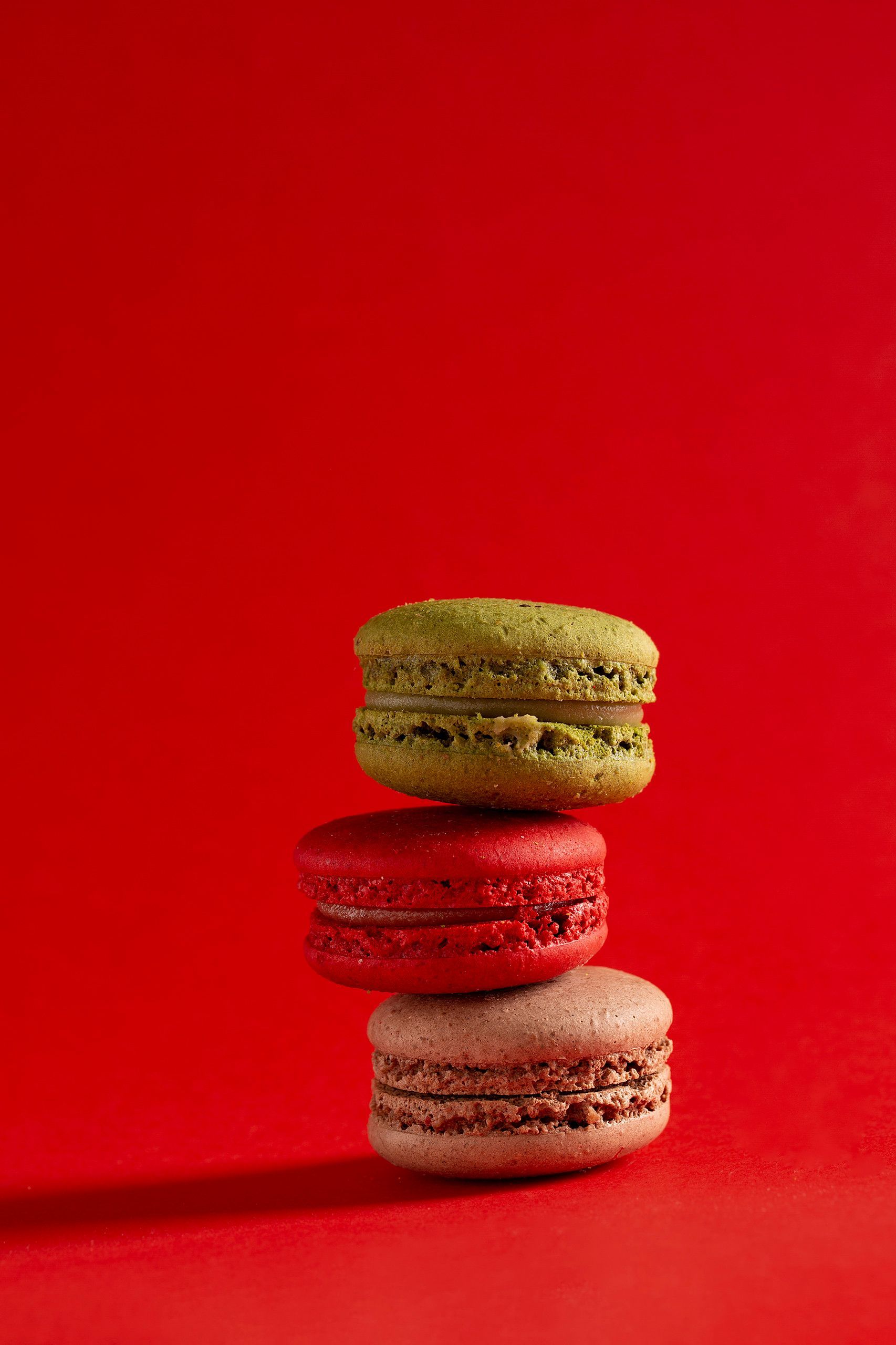 Três macarons empilhados nas cores verde, vermelha e rosa sobre um fundo vermelho vibrante.