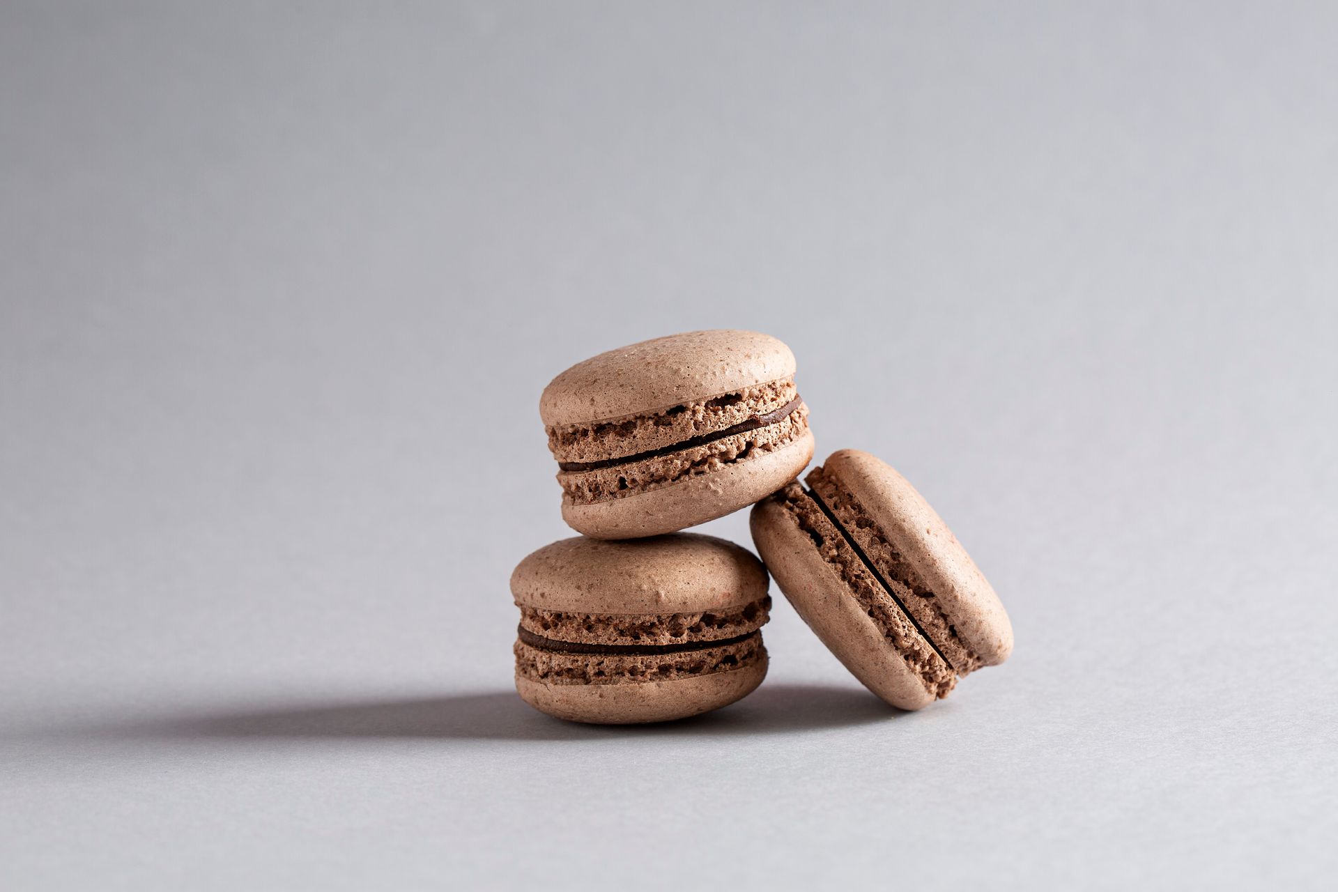 Três macarons bege empilhados sobre uma superfície cinza-clara, um encostado nos outros.