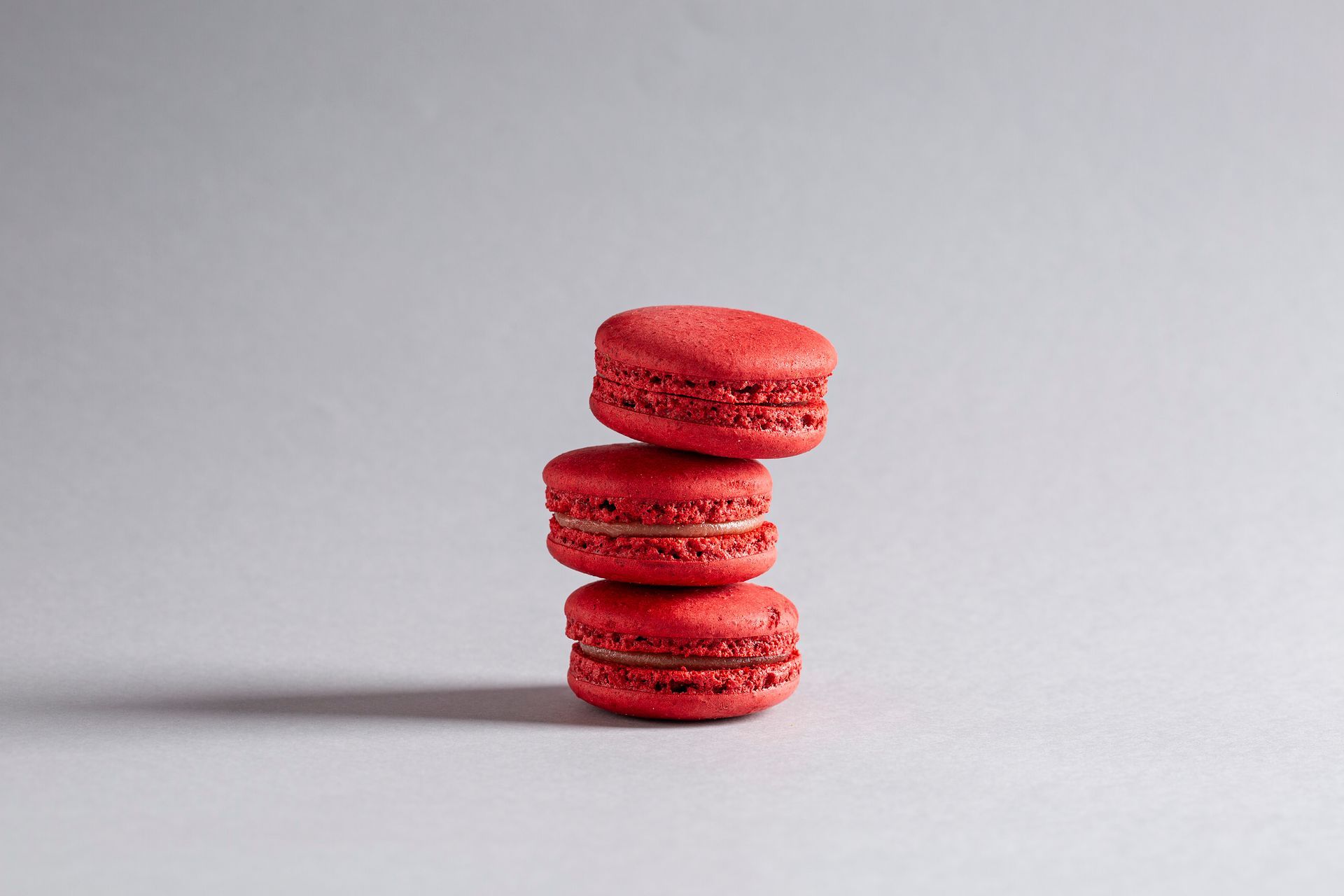 Três macarons vermelhos empilhados sobre uma superfície cinza-clara com uma sombra suave.