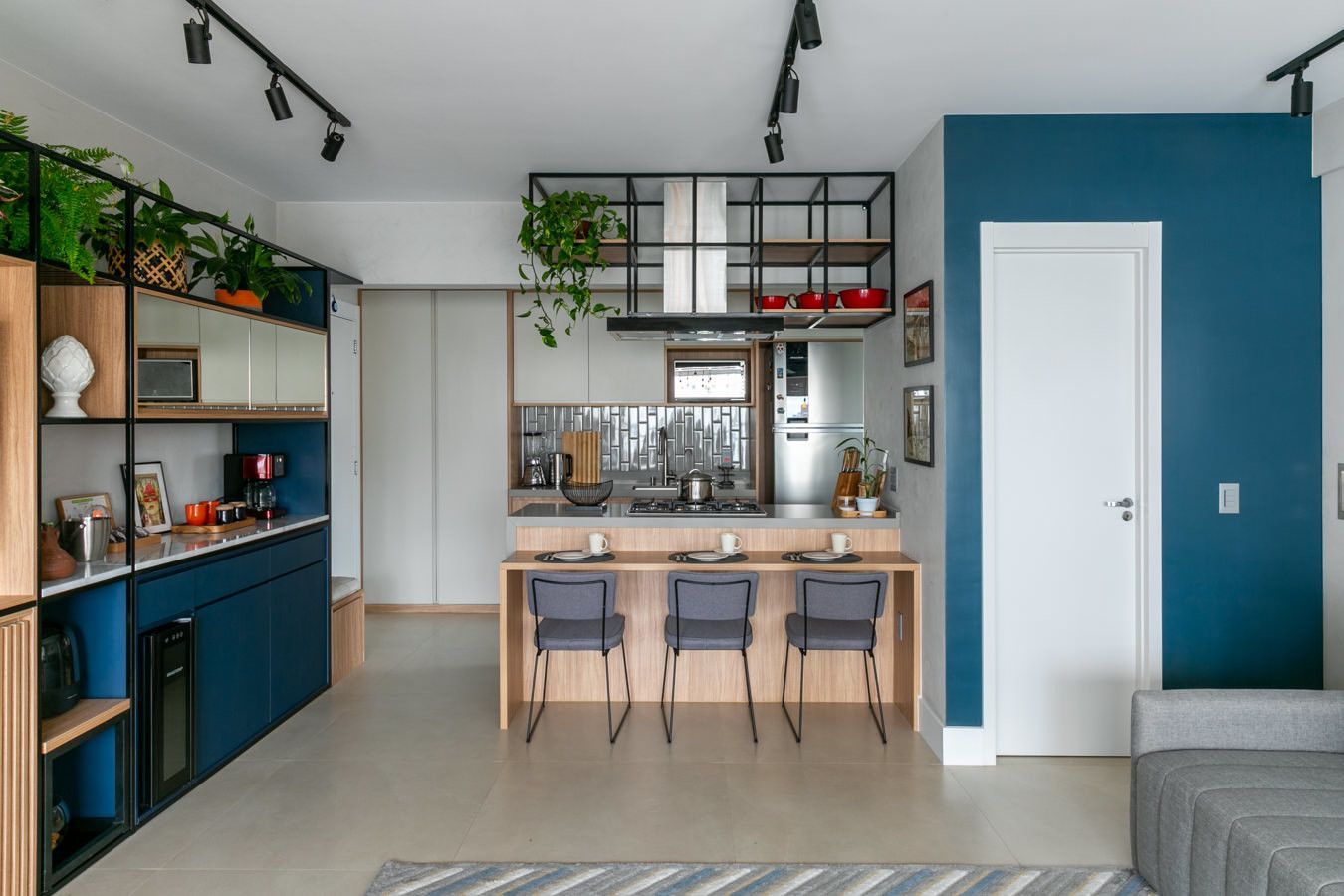 Cozinha e sala de estar modernas com ilha de madeira, parede de destaque azul, prateleiras pretas e plantas suspensas.