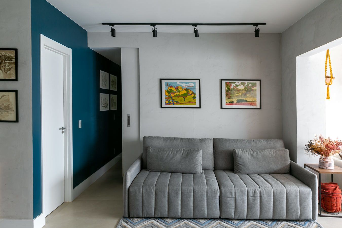 Sala de estar moderna com sofá cinza, parede de destaque azul, obras de arte emolduradas e iluminação em trilho.