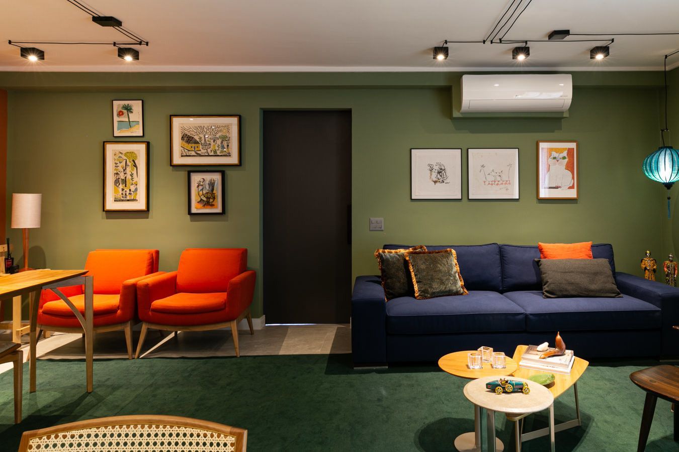 Sala de estar moderna com paredes verdes, sofá azul-marinho, cadeiras laranja, obras de arte emolduradas e mesa de centro sob iluminação direcionada.