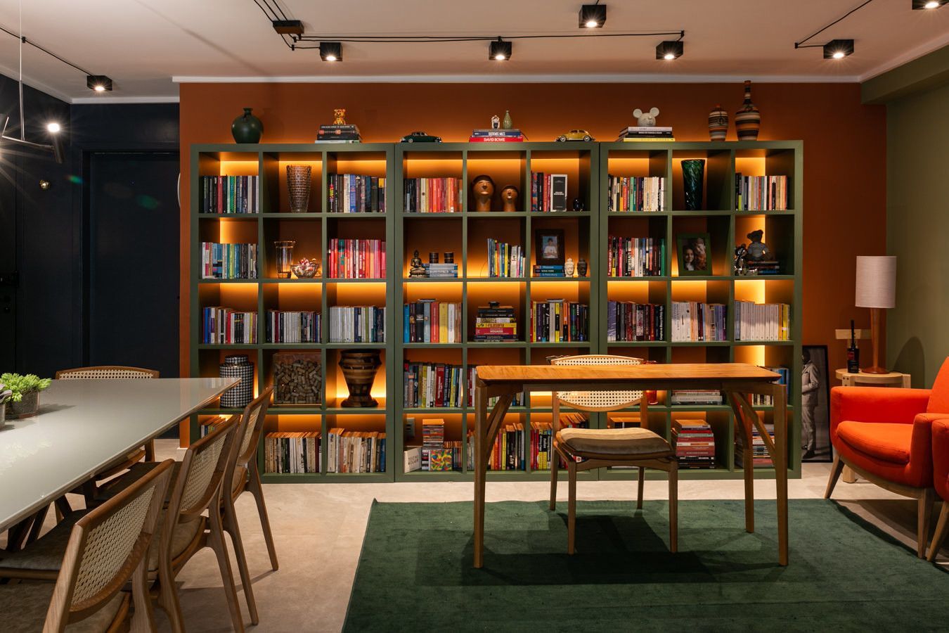 Sala de estar aconchegante com biblioteca, estantes do chão ao teto, mesa de madeira, cadeiras e assentos em um tom quente de laranja sob iluminação direcionada.