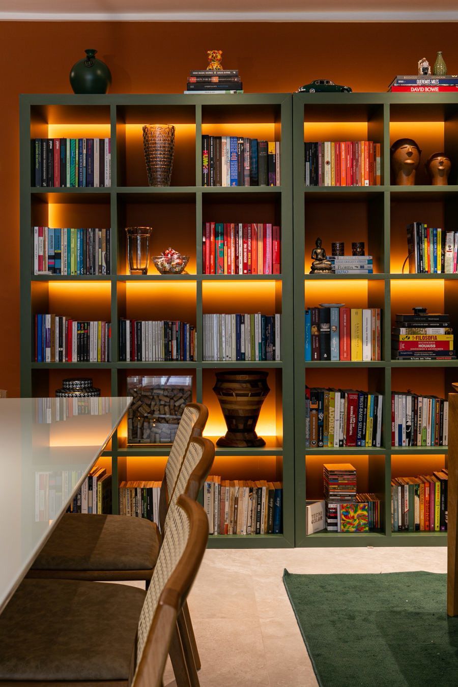 Parede com estante de livros iluminada com luz quente, livros coloridos, uma cadeira de jantar e uma mesa em um ambiente moderno.