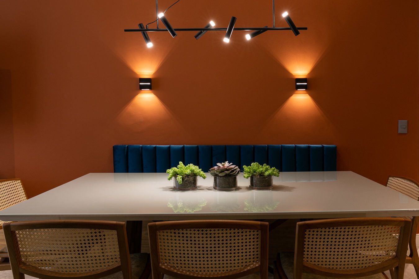 Sala de jantar moderna com paredes laranja, luminárias pendentes, mesa comprida e banco azul.