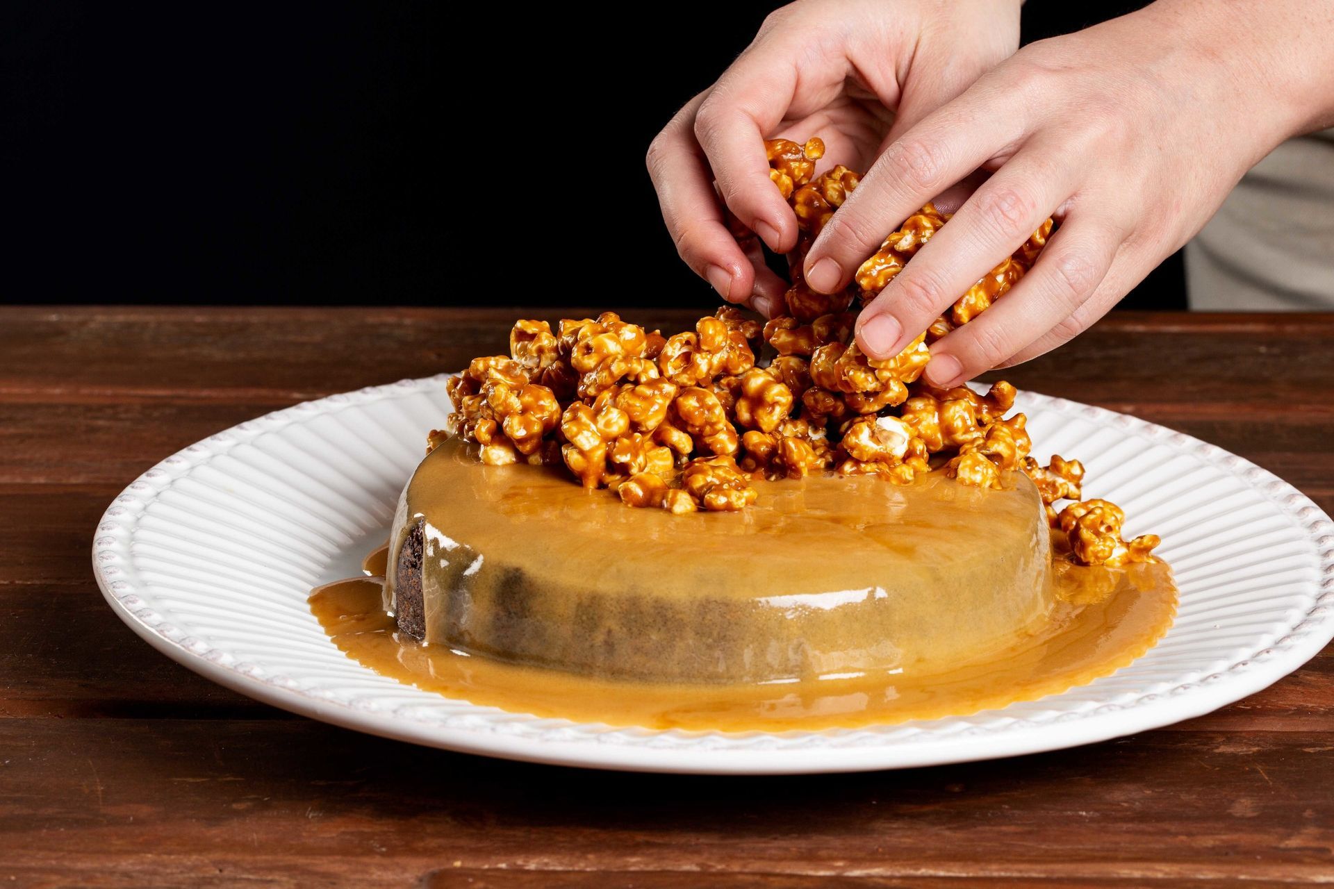 Mãos espalhando pipoca caramelizada sobre um bolo com cobertura de caramelo em um prato branco.