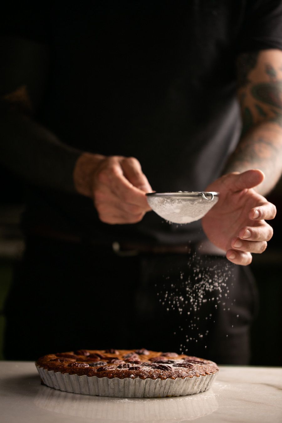 Uma pessoa polvilha açúcar de confeiteiro sobre uma torta em uma mesa em uma cozinha escura.
