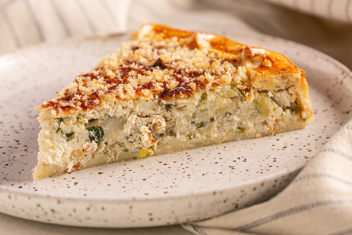 Fatia de quiche em um prato salpicado, coberta com queijo dourado e ervas.
