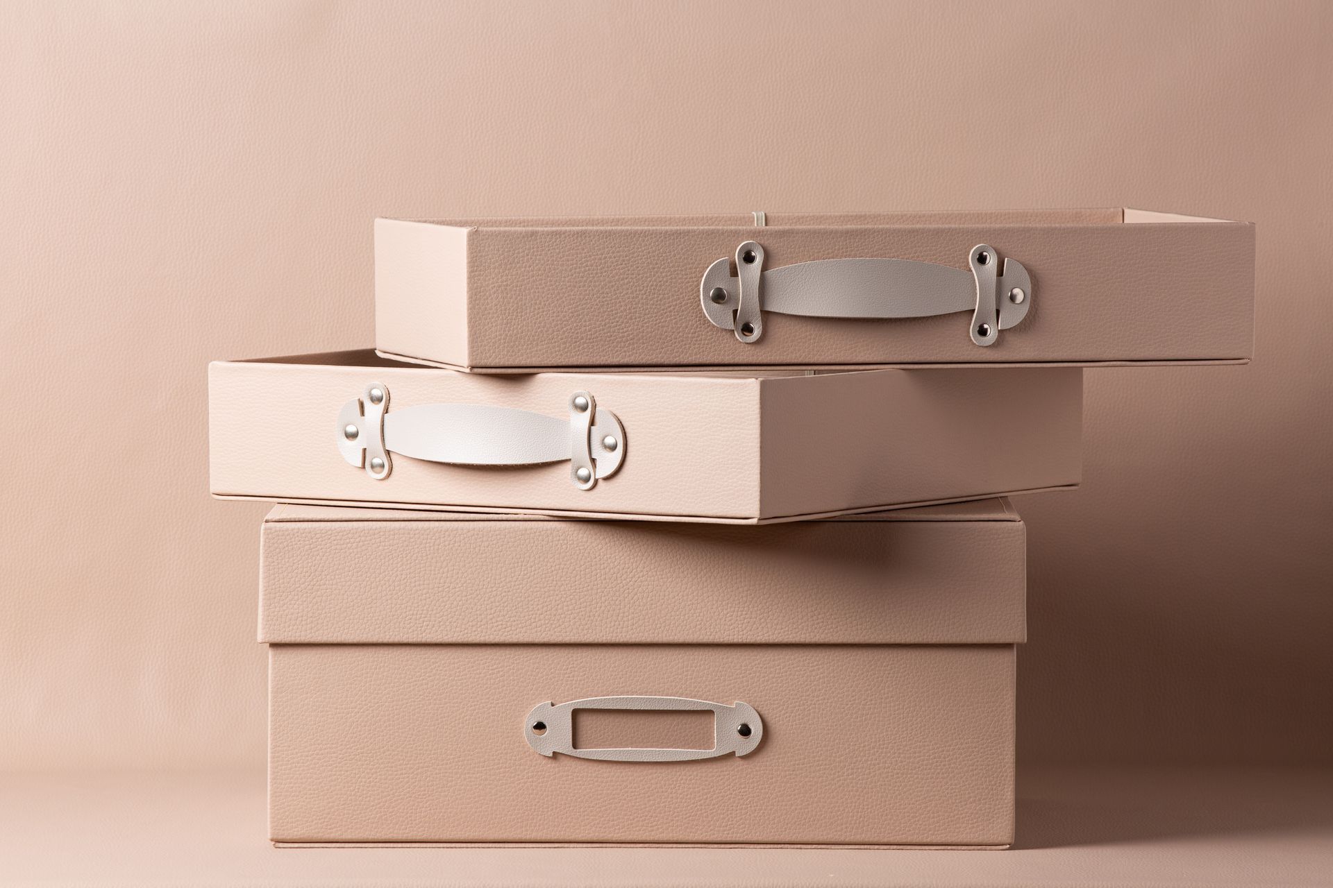 Caixas organizadoras bege empilhadas com alças de metal sobre um fundo rosa suave.