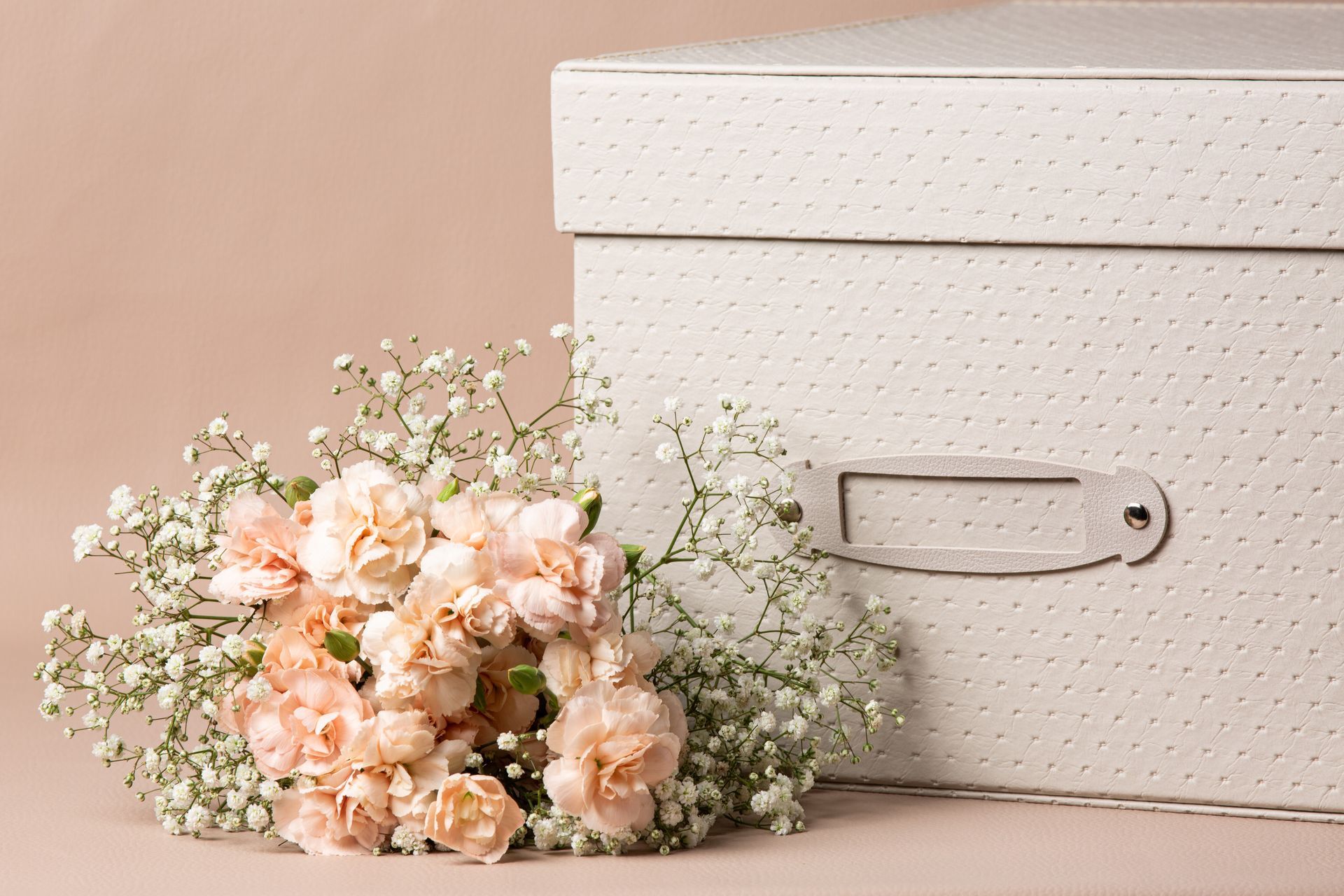 Caixa de armazenamento branca ao lado de um buquê de flores brancas e cor de pêssego sobre um fundo bege.