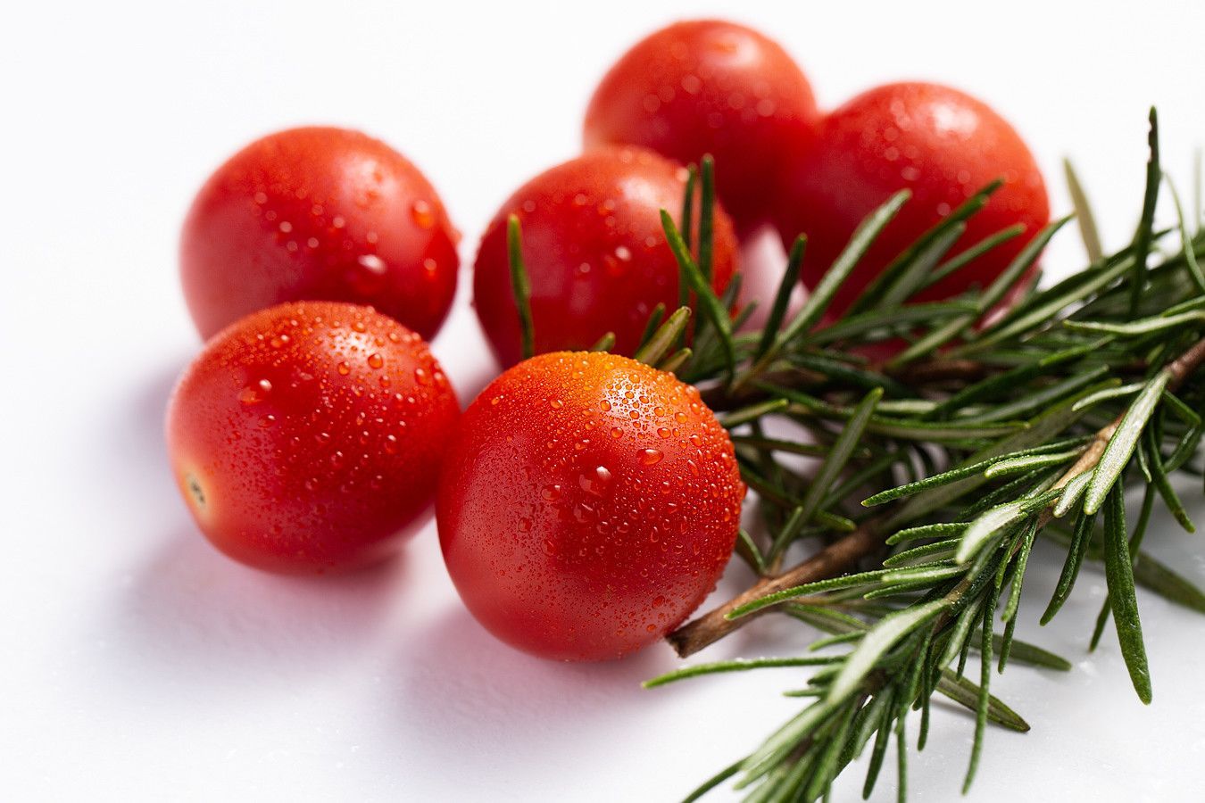 Tomates-cereja vermelhos com raminhos de alecrim sobre uma superfície branca.