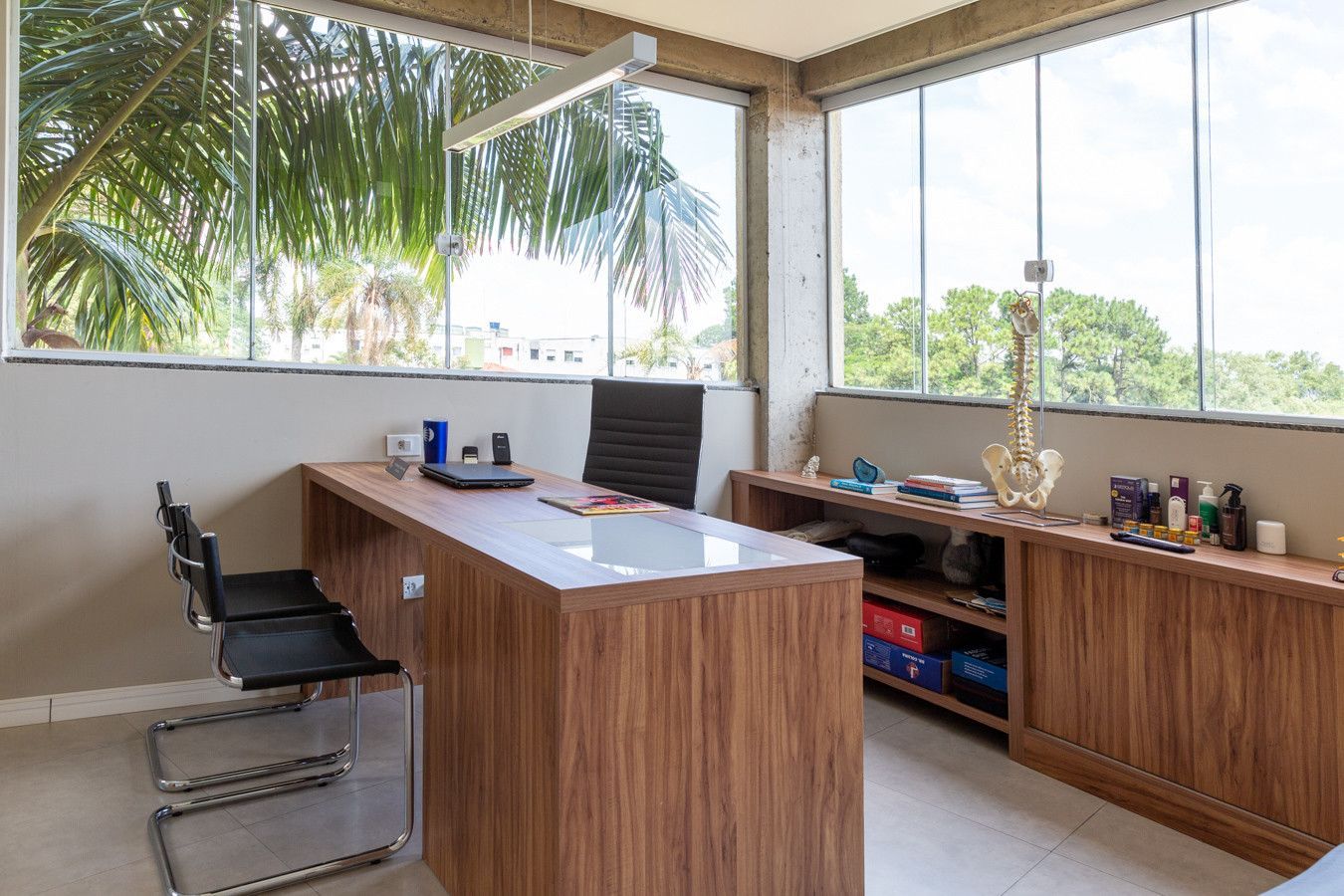 Escritório moderno com mesa de madeira, cadeiras pretas e grandes janelas com vista para palmeiras.