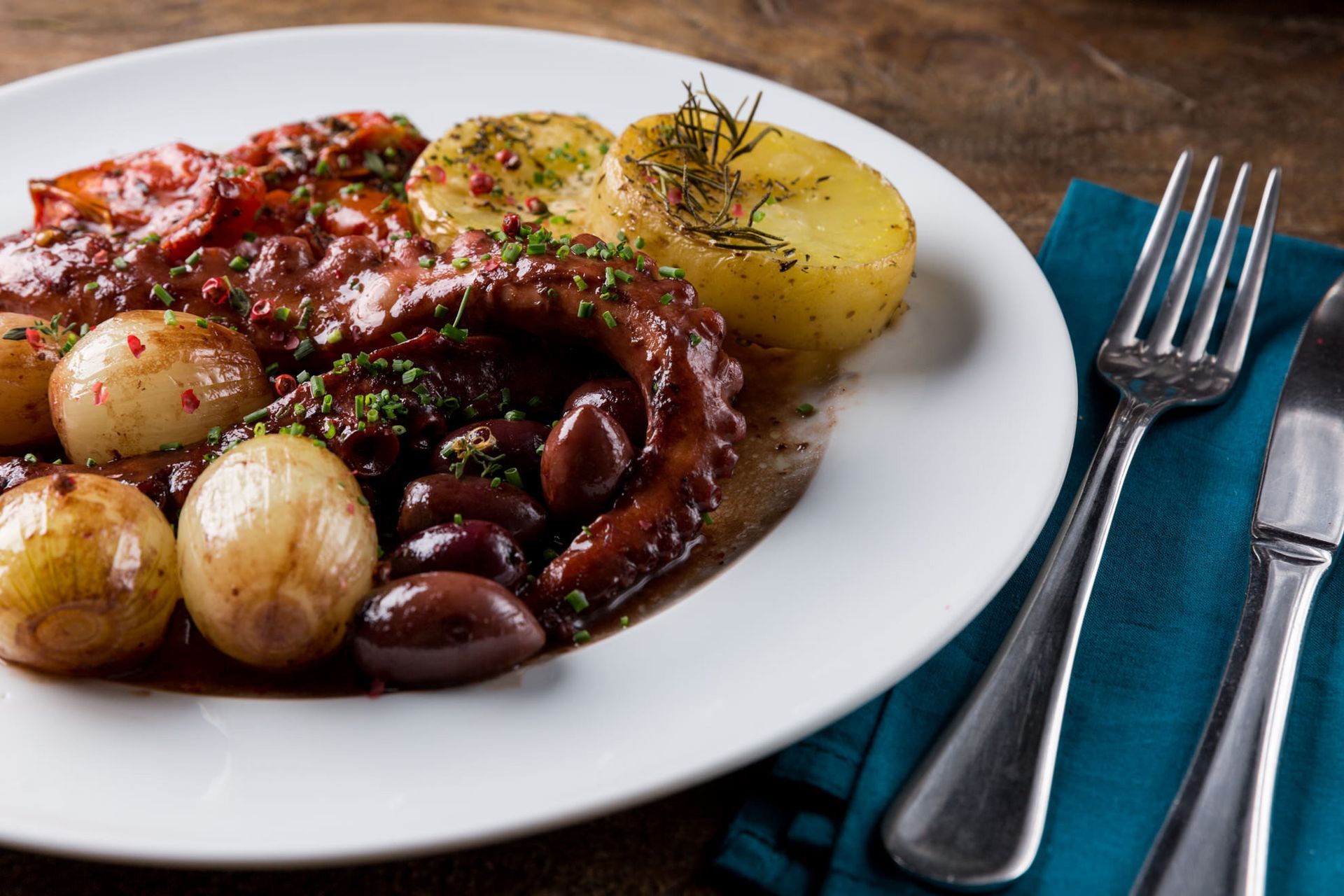 Prato de polvo e batatas ao molho vermelho com azeitonas, servido com garfo e faca sobre um guardanapo azul.