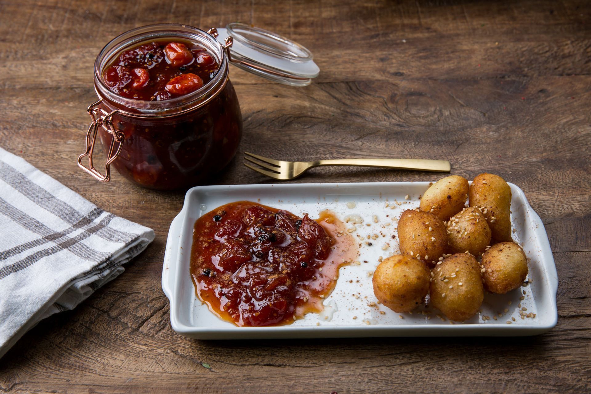 Pote de geleia de frutas vermelhas ao lado de um prato com geleia e bolinhos de massa fritos dourados em uma mesa rústica.