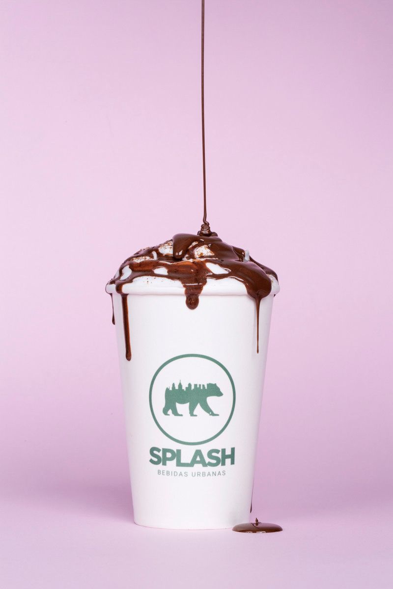 Xícara branca com o logotipo verde da Splash, coberta e com cobertura de chocolate escorrendo sobre um fundo rosa.