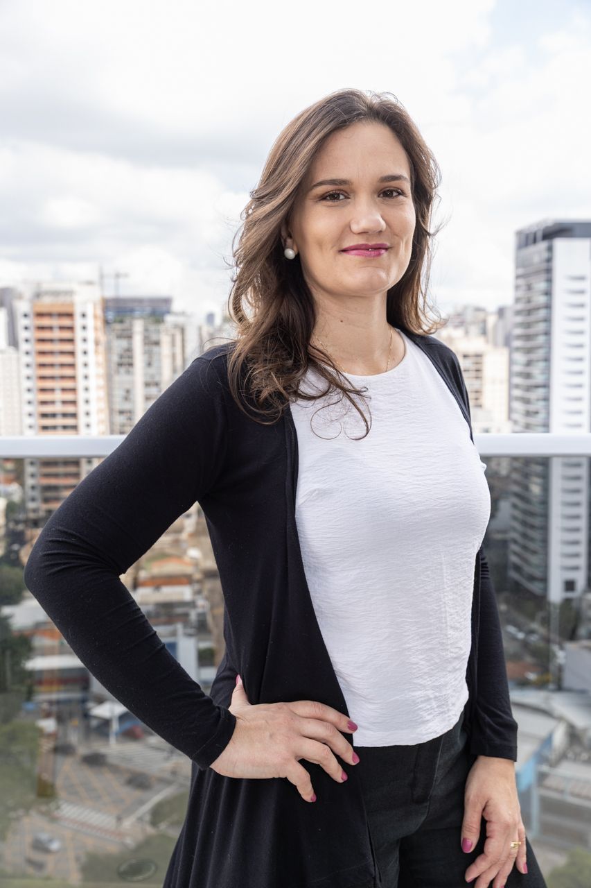 Mulher em pé numa varanda com prédios da cidade ao fundo, vestindo uma blusa branca e um cardigã preto.