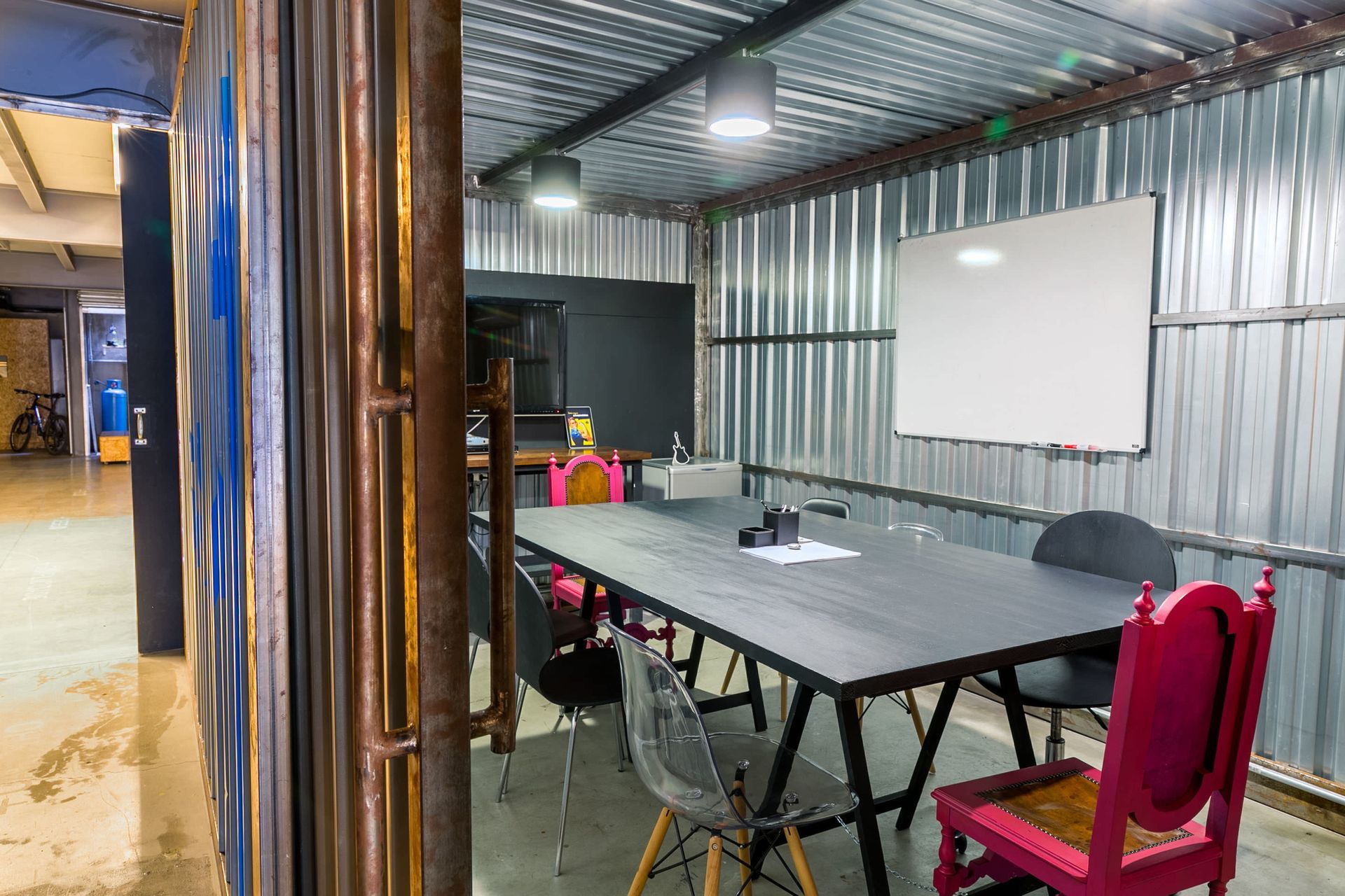 Sala de reuniões com mesa comprida, cadeira vermelha, tela de projeção e paredes de metal ondulado.