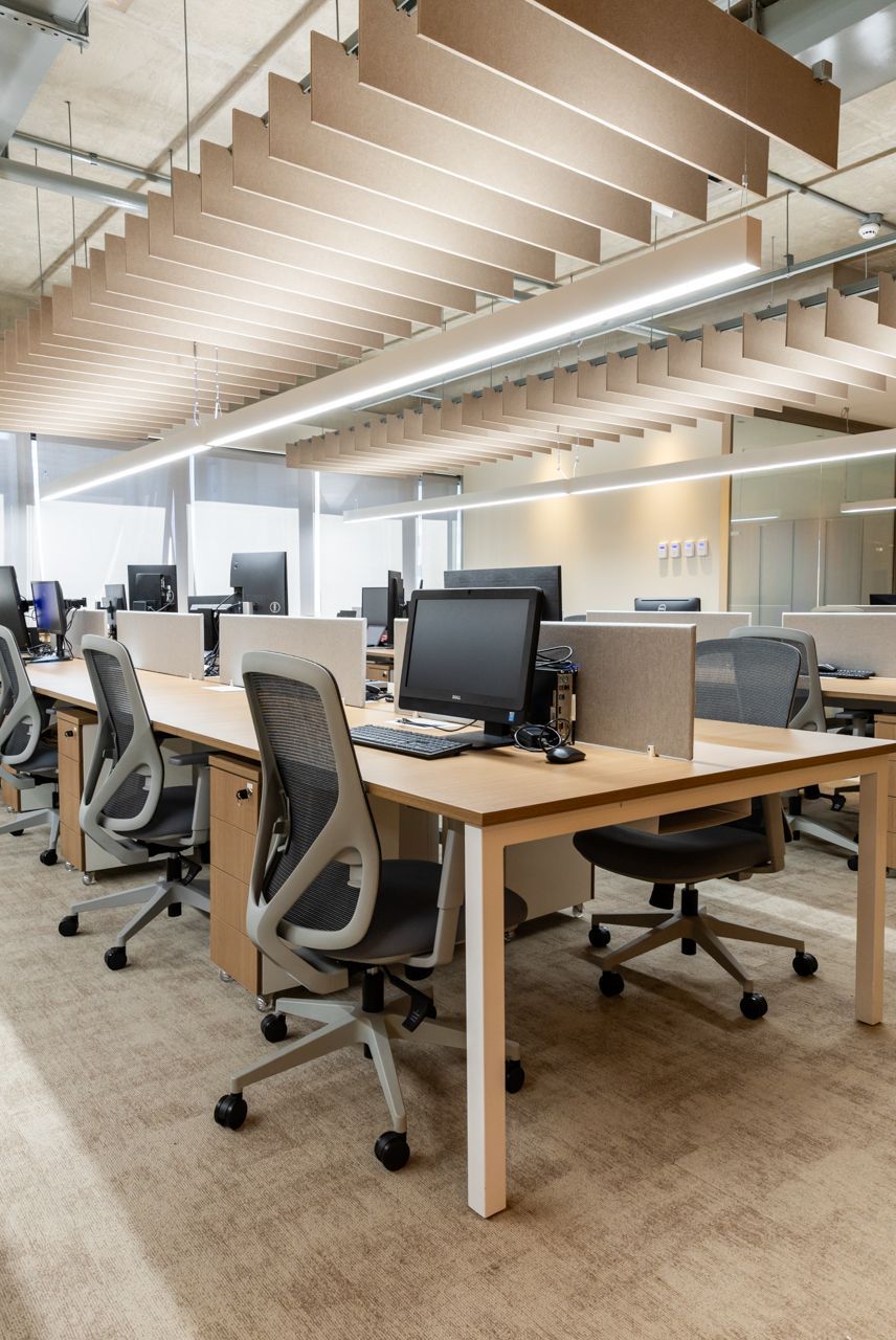 Escritório moderno e aberto com fileiras de mesas, monitores de computador e cadeiras ergonômicas sob um teto iluminado.