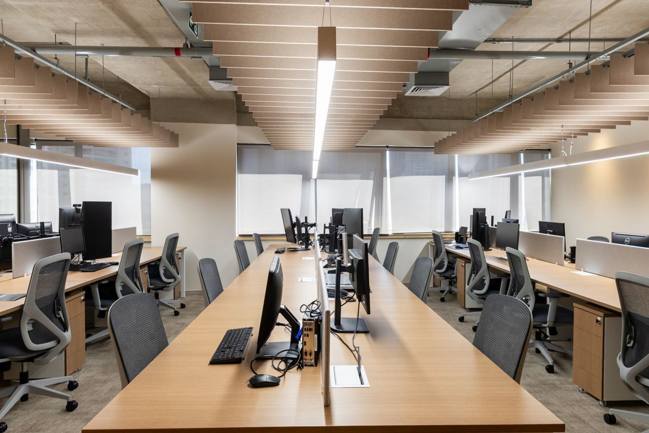 Escritório de planta aberta com fileiras de mesas, monitores e cadeiras cinza sob iluminação forte no teto.