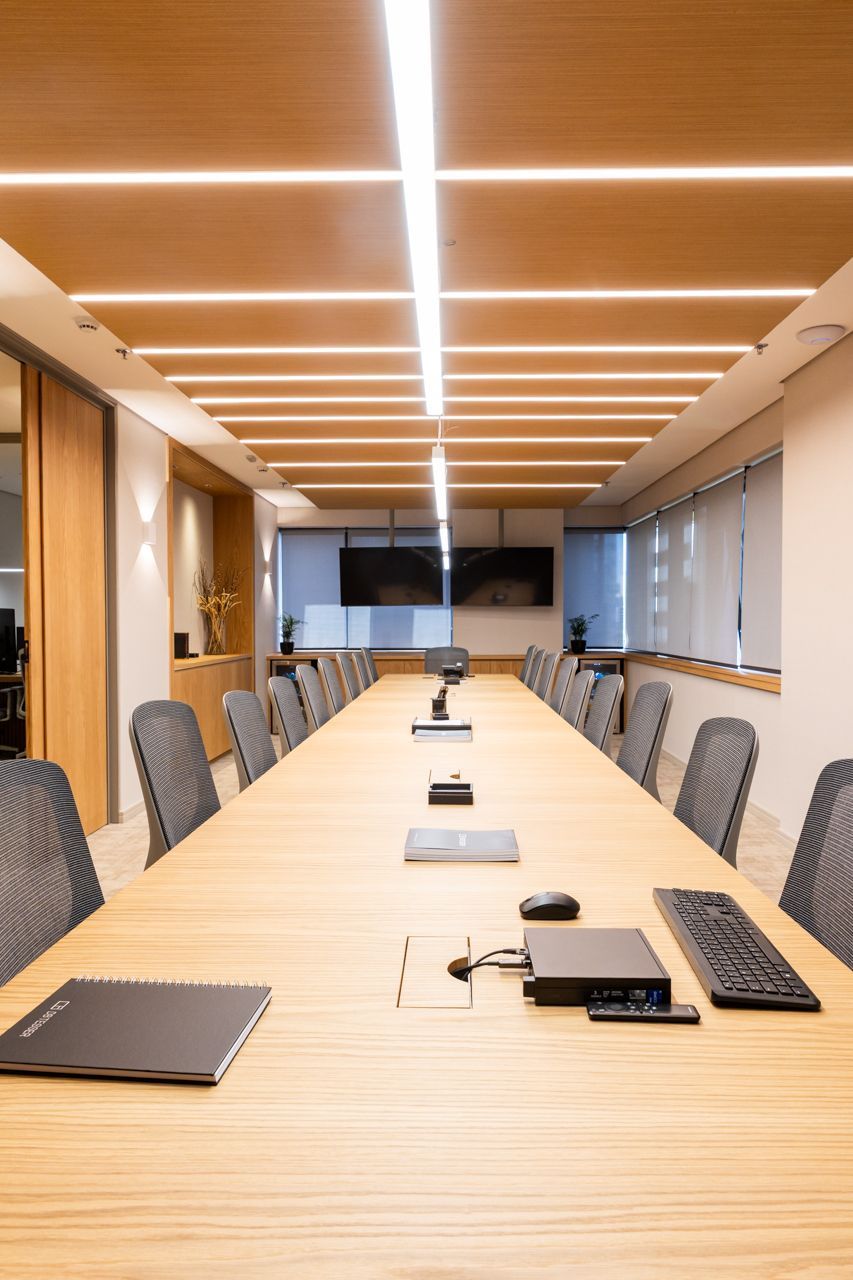 Sala de conferências moderna com uma longa mesa de madeira, cadeiras cinzentas e iluminação embutida no teto.