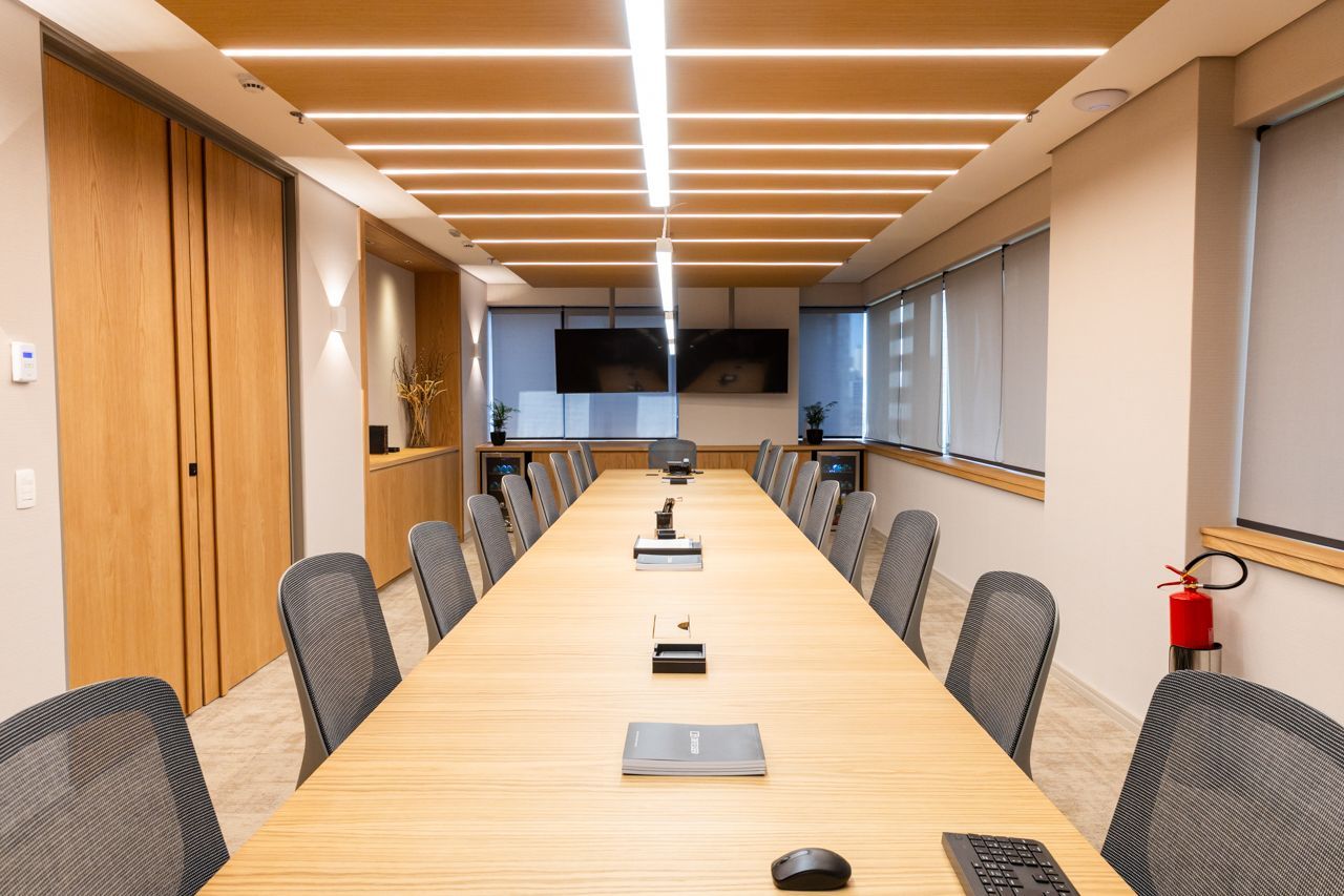 Sala de conferências moderna e vazia, com uma longa mesa de madeira, cadeiras cinzentas, divisória de parede e extintor de incêndio.