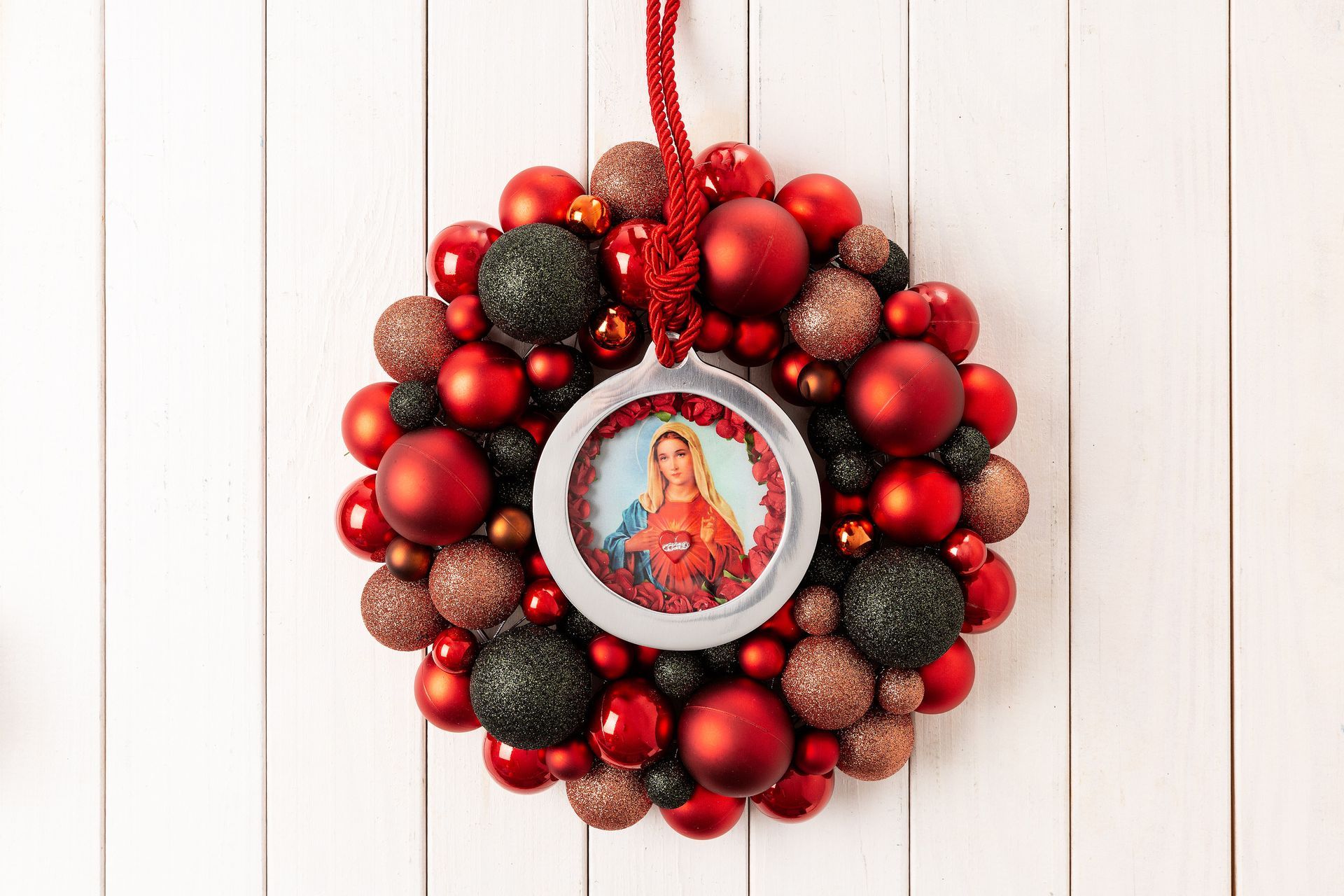 Coroa de flores vermelha com medalhão religioso central sobre fundo de madeira branca.