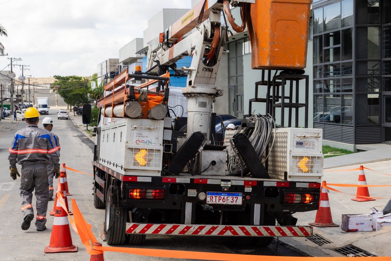 Operários de serviços públicos, usando capacetes de segurança, consertam uma rua a partir de um caminhão com plataforma elevatória, com cones de trânsito bloqueando a faixa.