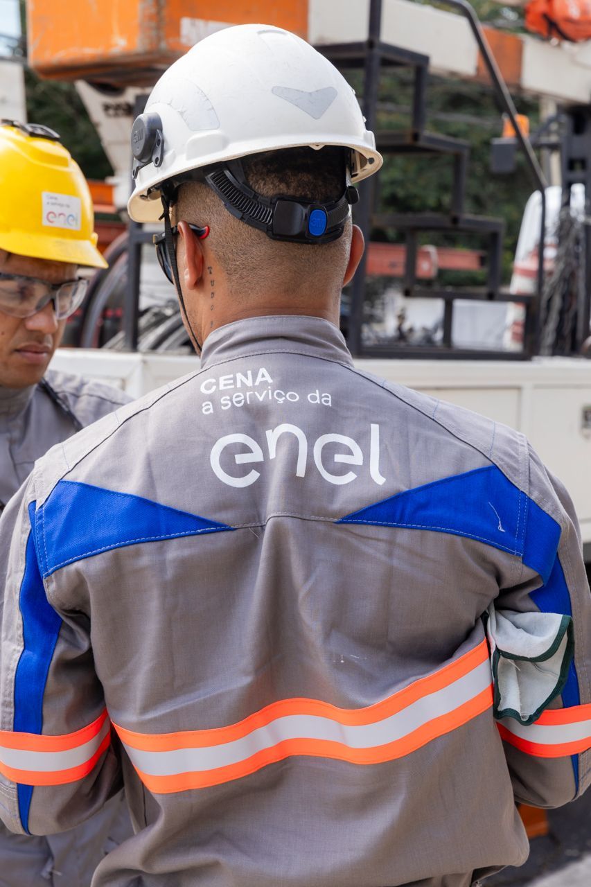 Operários com capacetes e coletes de segurança em um canteiro de obras, um deles com o logotipo da Enel nas costas.