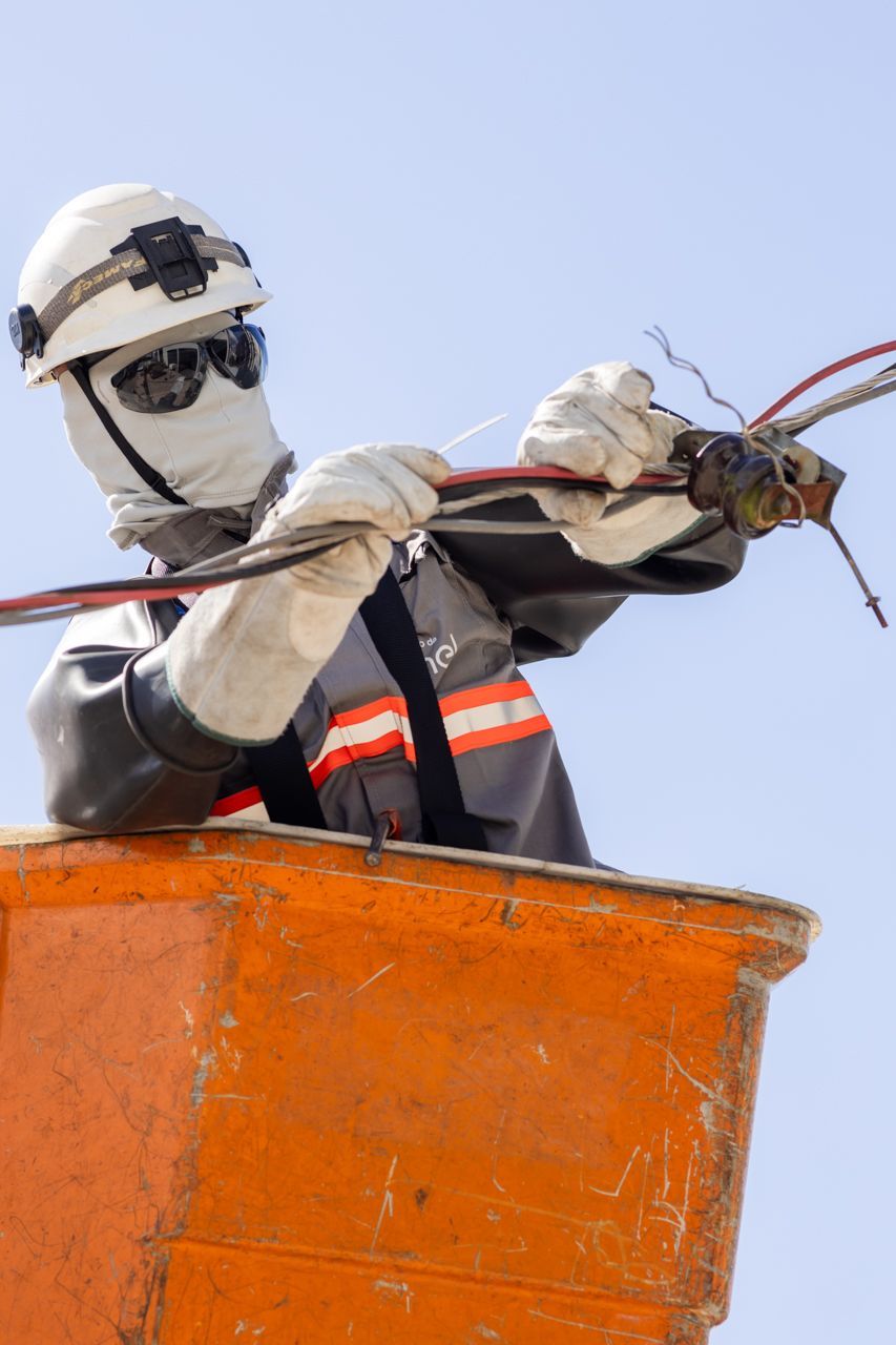 Operário de serviços públicos, com capacete branco e óculos de sol, manuseia cabos de uma plataforma elevatória laranja contra um céu azul.