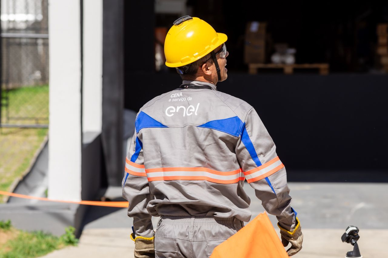 Operário com capacete amarelo e colete refletor cinza, em pé perto da entrada de um prédio.