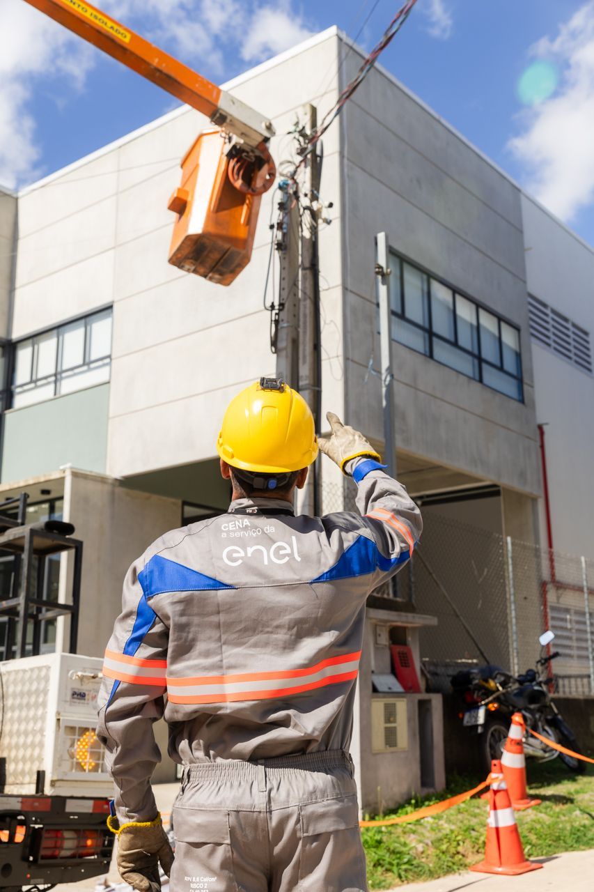 Operário de serviços gerais com capacete amarelo orientando uma plataforma elevatória ao lado de um prédio em manutenção.