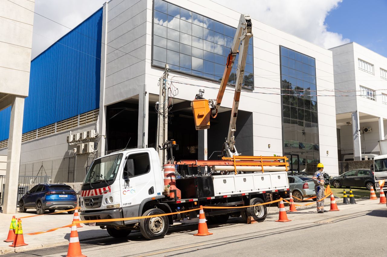 Caminhão de construção e guindaste estacionados ao lado de um prédio com operários, cones de sinalização laranja e barricadas em uma rua da cidade.