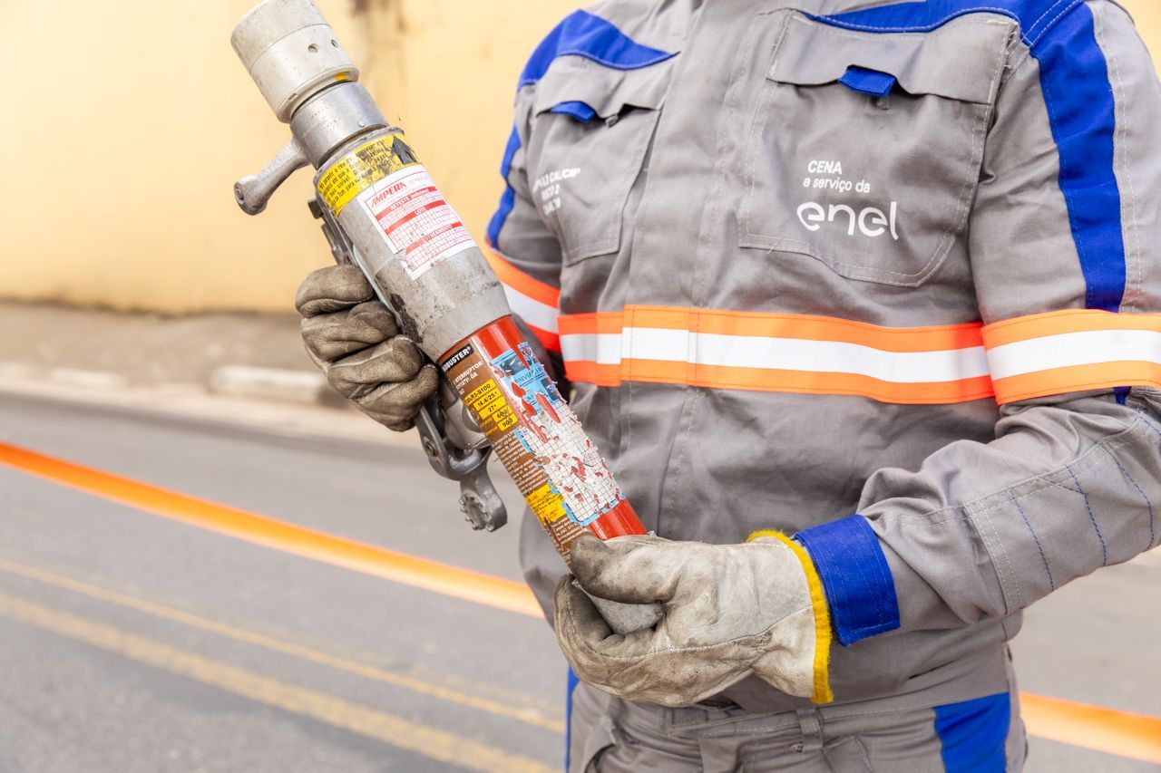 Trabalhador com uniforme cinza e azul segurando um extintor de incêndio com listras vermelhas à beira da estrada.
